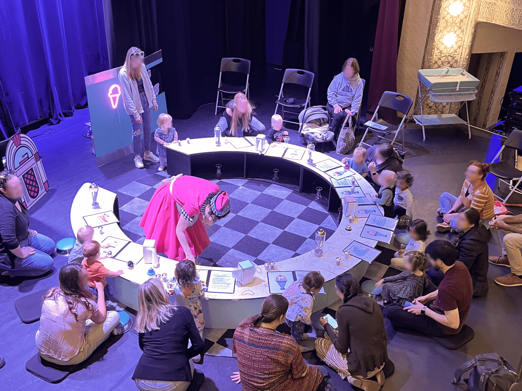 Babies, toddlers and their caregivers sit around a circular table in a purple-lit space. The main performer in the interactive show bends down to one of the audience members.